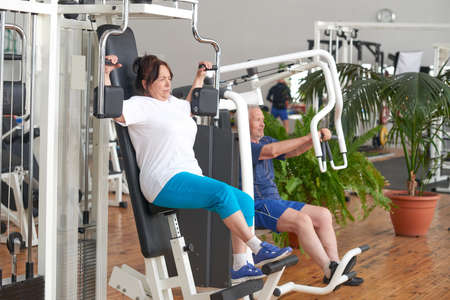 Senior woman training on machine at gym. Elderly people exercising at fitness club. How to help...