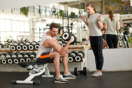 Sportive man flexing muscles with dumbbell. Delighted woman looking on strong man exercising with...