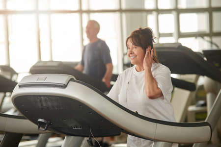 Senior woman on treadmill. Pretty elderly woman listening to music during workout at gym. Sport as a...