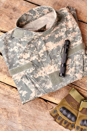 Set of military outfit on brown wooden background, flat lay. Folded camouflage, torch and fingerless...