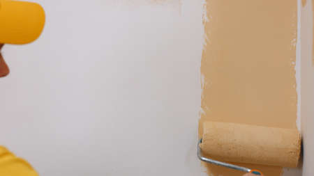 Workman painting wall with brush paint roller. Man with paint roller painting white wall at interior using beige color.の写真素材