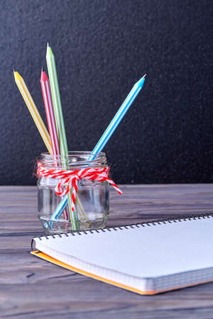 Composition with school supplies on wooden background. Blank notebook and jar with pencils on table....