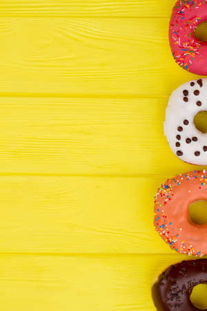 Tasty donuts with colorful sprinkles, cropped image. Border from different glazed donuts on yellow wooden background. Space for text.の写真素材