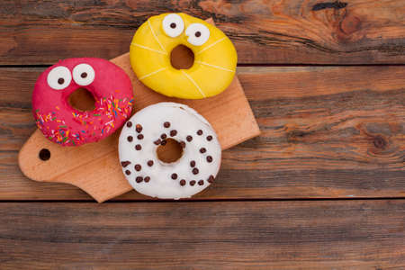 Funny decorated donuts on wooden background. Three different donuts with sugar icing on cutting board. Flat lay, top view.の写真素材