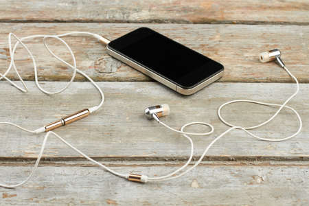 Smartphone with earphones on wooden boards. Mobile phone with blank screen and earbuds on vintage wooden background. Modern audio technology.の写真素材