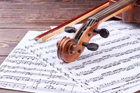 Violin scroll and peg box on musical notes. Music note sheets, violin and fiddle stick on wooden background. Detail of violin. Classical music concept.の写真素材