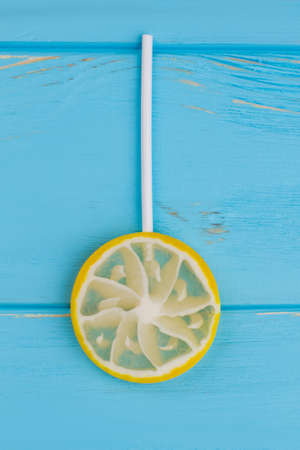 Candy lemon slice on stick. Yellow lollipop in a shape of lemon on blue wooden background, top view. Minimal summer concept.の写真素材