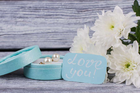 Flowers and gift box with jewelry. Present box with pearl earring, bouquet of chrysanthemums and card with inscription love you on wooden background. Happy Valentines Day.の写真素材