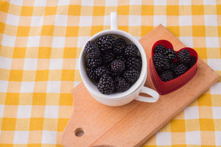 Cup and heart-shaped form are filled with blackberries. Wooden kitchen board, textile checkered...