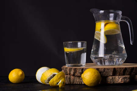 Lemons around wooden cut. Jug and glass of lemon water. Black studio.