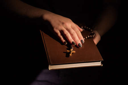 Womans holding holy bible with both hands. Wooden rosary with crucifix, black background.