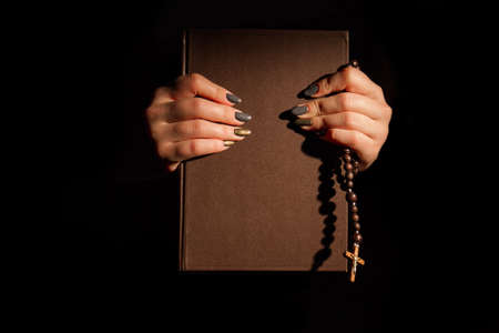 Woman holds brown holy book bible and rosary. Dark black background.