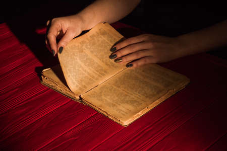 Female hand turning page of very old ancient book. Red wooden table surface.の写真素材