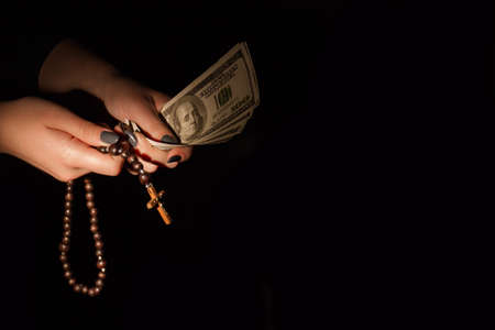 Money and religion concept. Closeup young female hands with dollars currency and wooden cross on...
