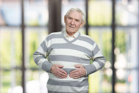 Senior caucasian man suffers from stomachache. Portrait of old grandfather feeling pain in his stomach. Blurred windows background.の写真素材