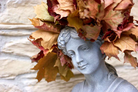 Stone head of a young female sculpture with autumn leaves wreath. Face close up.