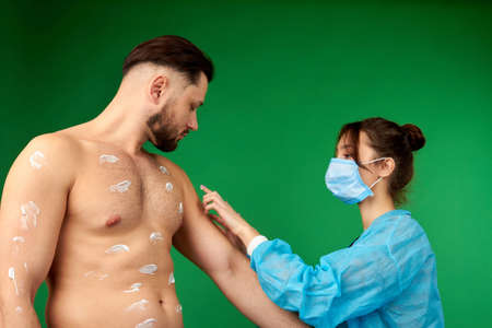Doctor making ointment spots on her patients body. Strong young man with white smears on his body. Green isolated background.の写真素材