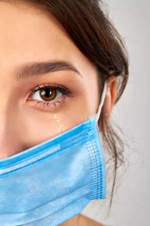 Close up crying womans eye. Wearing protective blue medical mask. Isolated on grey background.