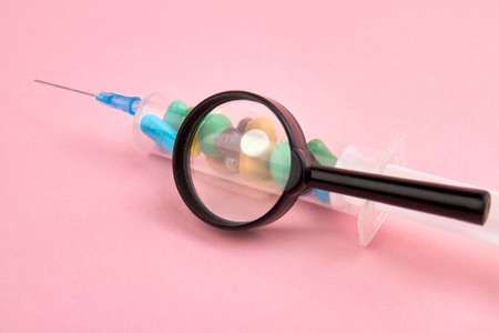 Syringe filled with medicine pills and glass magnifier. Isolated on pink background.