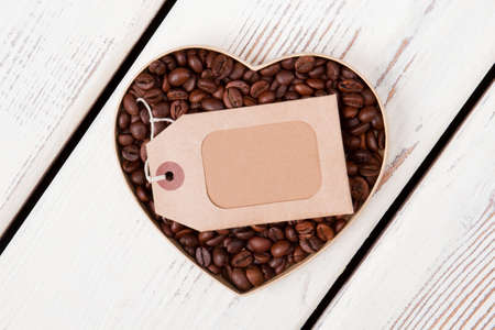 Heart shaped coffee beans with blank paper tag. Label for copyspace. White wooden background.の写真素材