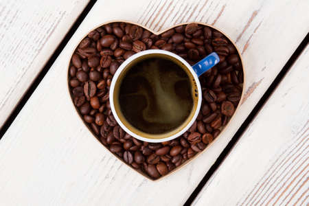 Cup of coffee standing over heart made of coffee grains. Coffee love concept. White wooden...