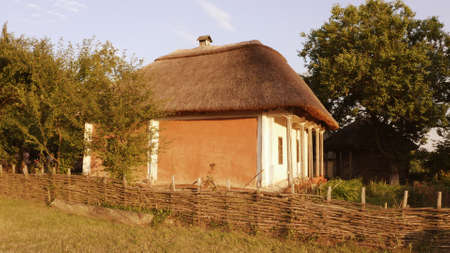 Old east european rural wooden house with wattle fence and thatched roof. Sunny summer day.の写真素材