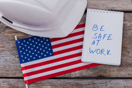 Flat lay warning american builders workers to be safe at work. White helmet with flag of the USA and notepad on aged wooden table.の写真素材
