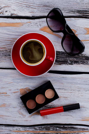 Cup of coffee with sunglasses and makeup accessories. Mascara and powder. White vintage wooden background.の写真素材