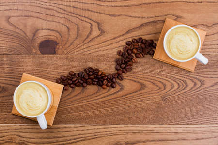 Two cups of coffee cappuccino conncted with coffee beans. Topview flat lay. Brown wooden table surface on the background.の写真素材