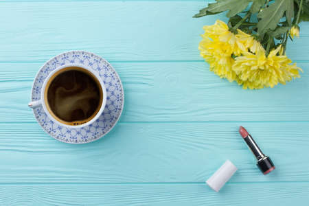 Flat lay cup of coffee with flowers and lipstic. Copy space and blue wooden table on the background.