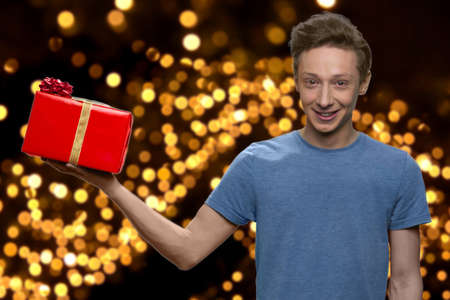 Teen boy holding gift box on his palm. Golden bokeh lights on black background. Winter holiday...