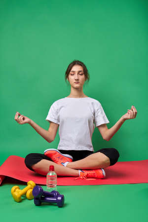 Young girl with closed eyes in meditation pose. Relaxation yoga pose concept. American girl in white shirt is meditating on red yoga mate. Isolated on green background.の写真素材