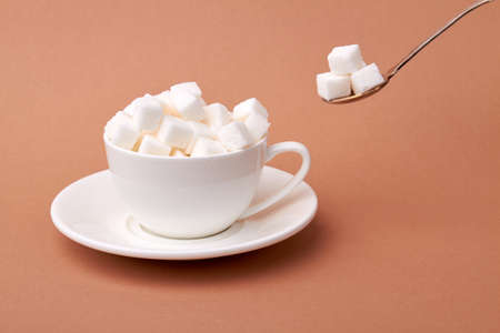 White cup of coffee is overfilled with sugar cubes. Too sweet beverage concept. Isolated on brown...