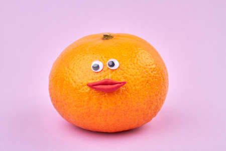 Ripe orange with funny female face. Citrus fruit with red womans eyes and lips isolated on purple background.の写真素材