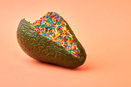 Avocado filling with sweet candy sprinklers. Close up green avocado full of multicolored sprinklers isolated on orange background.の写真素材