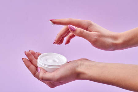Close-up female hand applying skin cream. Holding opened skicare cream jar isolated on purple background.の写真素材