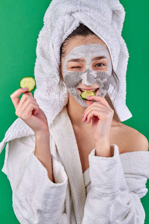 Girl with facial clay mask biting cucumber slice. Portrait of young woman wearing bathrobe and towel on her head.の写真素材
