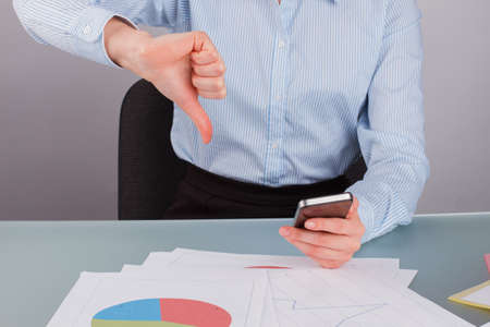 Close up businesswoman showing thumb down while working in office. Business lady holding smartphone...
