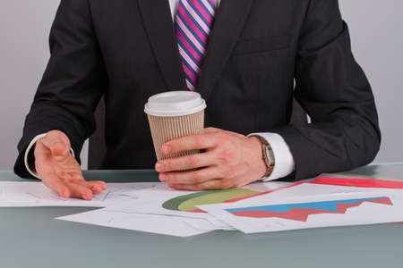 Business advisor analyzing company financial report. Businessman holding disposable coffee cup while working with documents at desk in office.の写真素材