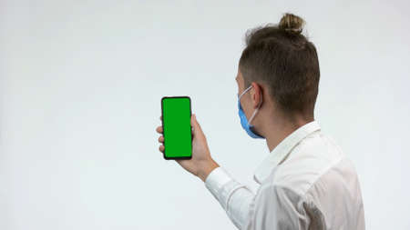 Man holds smarphone with green screen for copy space. Back view businessman with face mask holding smartphone with green screen. Isolated on white background.の写真素材