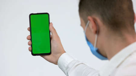 Man in face mask looks at green phone screen. Back view man wearing protective medical mask and looks at the green empty smarthone screen. Chromakey, copy space.の写真素材