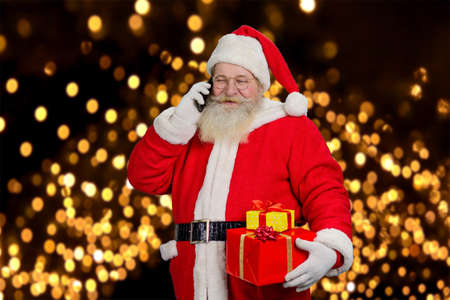 Santa Claus having Christmas call. Happy Santa Claus holding present boxes and talking on mobile phone on festive lights background.の写真素材