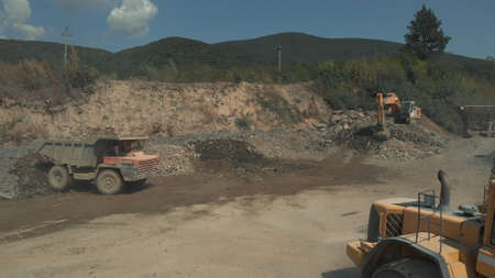 Bulldozer picks up stones. Trucks in the mountain carrier. Green hills on the background.