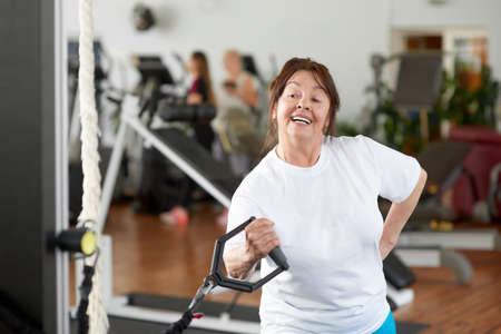 Pretty elderly woman working out at gym. Woman performs exercises with fitness trx system, TRX suspension straps. Physical hands endurance.の写真素材