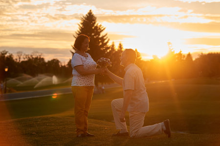 Man kneeling and giving flowers to his wife. Birthday or anniversary gift. Outdoors bright senset light.の写真素材