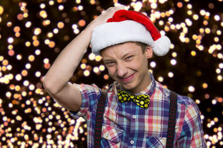 Cute caucasian teen boy in santas hat. Teenagers screwing up his eyes. Glowing lights on the background.の写真素材