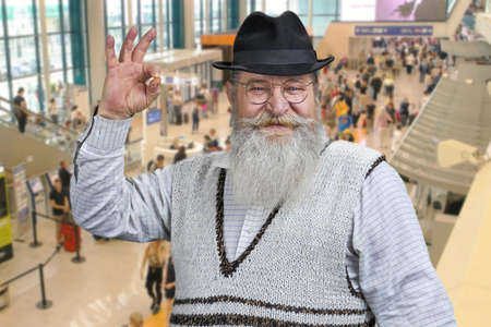 Happy senior man with beard gesturing okey while standing on escalator at airport.