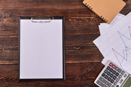 Clipboard with blank white paper and calculator. Brown wooden desk background.