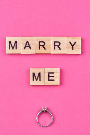 Flat lay top view of engagement ring and text marry me on wooden cubes. Proposal message on pink background.の写真素材