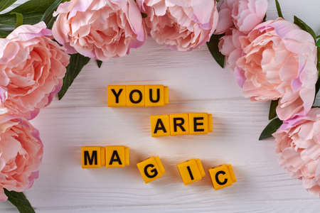Yellow cubes arranged in you are magic words. Frame of peonies on white desk.